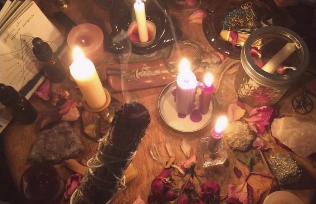 A candlelit altar with dried herbs, crystals, rose petals and a sage bundle